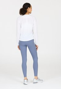 Vit långärmad topp med raglanärmar, kombinerad med åtsittande ljusblå leggings. Modellen bär silvriga sportskor. Slät textur genomgående.