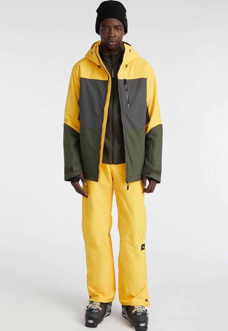 Mężczyzna ubrany w żółtą, szaro-zieloną kurtkę narciarską, żółte spodnie narciarskie, czarne buty narciarskie i czarną czapkę, stojący na tle jednolitego białego tła.
