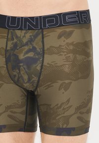 Camouflage-patterned boxer briefs in olive green with a black waistband featuring "UNDER" in gray. Material has a textured finish.