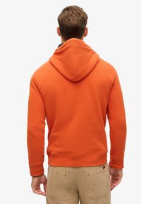 Hombre de pie de espaldas con sudadera con capucha naranja y pantalones beige sobre un fondo blanco.