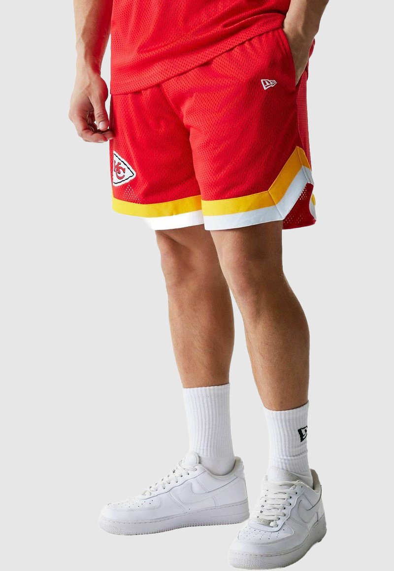 Shorts de sport rouges avec des accents jaunes et blancs, arborant un écusson, en tissu mesh, et une coupe décontractée, portés avec des baskets et des chaussettes blanches.