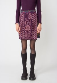 Short purple skirt with pink floral and geometric patterns, featuring a decorative waistband. Worn with black knee-high textured boots.