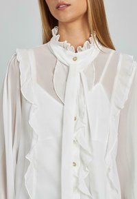 Weiße Bluse aus transparentem Stoff mit einem Rüschkragen, Knopfverschluss und zarten Rüschendetails entlang der Vorderseite und Ärmel.