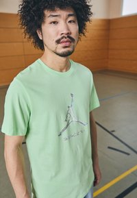 T-shirt in cotone verde chiaro con una grafica argentata di una figura che salta e la parola "SPORT" sottostante. Maniche corte e vestibilità classica.