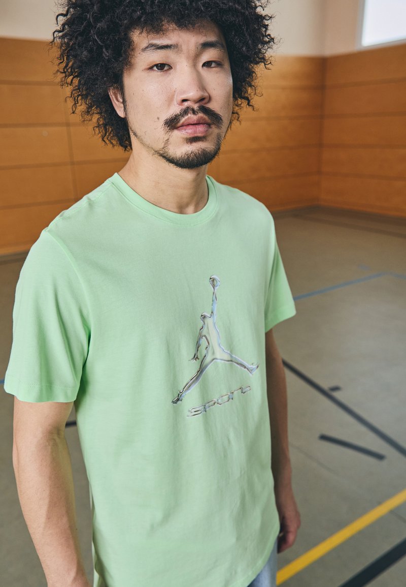 T-shirt in cotone verde chiaro con una grafica argentata di una figura che salta e la parola "SPORT" sottostante. Maniche corte e vestibilità classica.