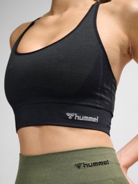 Femme portant un brassière de sport noire et des leggings taille haute vert olive, tous deux avec le logo hummel, montrée du ventre aux épaules.