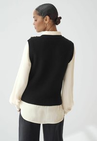 Gilet en tricot noir avec un motif texturé, présentant un col rond et sans manches, superposé sur une chemise blanche à manches longues.