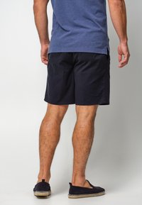 Shorts de color navy hechos de algodón, con un corte relajado, largo hasta la rodilla y combinados con zapatos oscuros de tipo slip-on sobre un fondo claro.