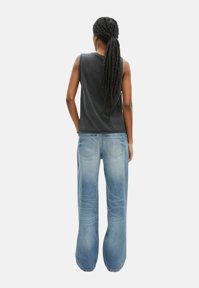 Zwarte mouwloze top, blauwe wijde spijkerbroek. De denim heeft een vervaagde wassing met subtiele diagonale lijnen. De achterzakken zijn zichtbaar. Lange gevlochten haarstijl.