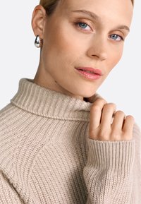 Beige gerippter Strickpullover mit hohem Kragen und umgeschlagenen Ärmeln, der ein strukturiertes Muster und eine lockere Passform aufweist. Sichtbares Detail mit silbernen Ohrringen.