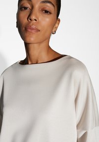 Frau mit glatter Haut, die silberne Creolen trägt und ein lockeres, strukturiertes weißes Oberteil vor einem schlichten weißen Hintergrund trägt.