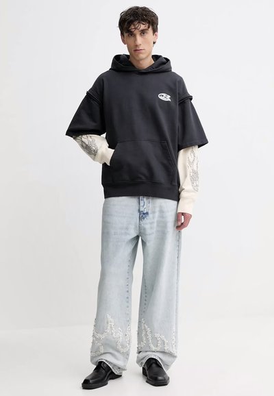 Felpa nera oversize con tasca frontale, maniche color crema con motivo floreale, abbinata a jeans a gamba larga di un blu chiaro con orli decorati.