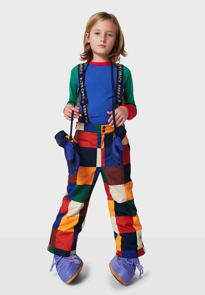 Farverigt ski-outfit med en blå langærmet trøje og multifarvede ternede bukser, der har stropper og blå vanter. Lilla støvler fuldender looket.