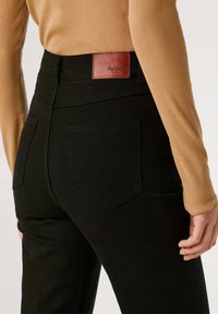 Jeans negros de cintura alta con un parche de cuero de la marca, que presentan dos bolsillos traseros y una textura suave. Llevados con una blusa de manga larga beige.