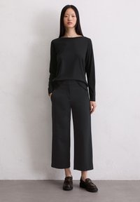 Crna majica dugih rukava, široke crne hlače i tamne mokasine. Glatke teksture s elegantnim dizajnom. Neutralna pozadina.