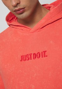 Felpa capuccio corallo realizzata in morbido tessuto, caratterizzata da ricamo tonale "JUST DO IT." sul davanti, con una vestibilità comoda e una finitura testurizzata.