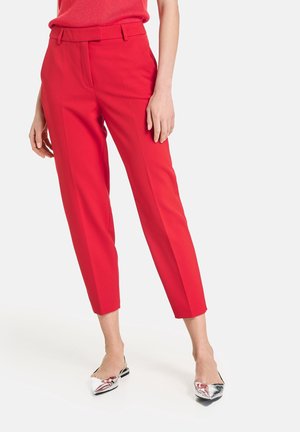 Femme portant un pantalon rouge vif ajusté à la cheville et des chaussures slingback argentées à bout pointu, debout devant un fond uni.