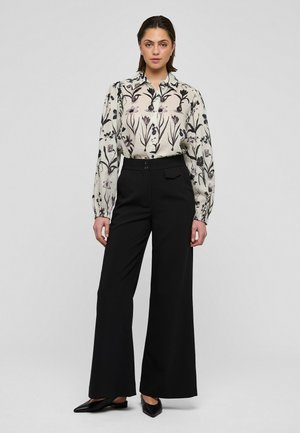 Ženska, ki nosi široke črne hlače, črne koničaste čevlje in belo bluzo s črnimi cvetličnimi vzorci, stoji ob svetlem enobarvnem ozadju.