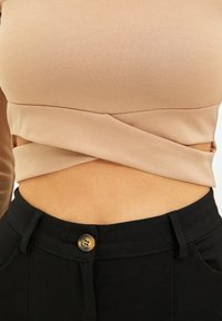 Tailliertes hellbraunes Oberteil mit einem strukturierten, gedrehten Design an der Taille, kombiniert mit hochgeschnittenen schwarzen Hosen, die einen glänzenden goldenen Knopf haben.