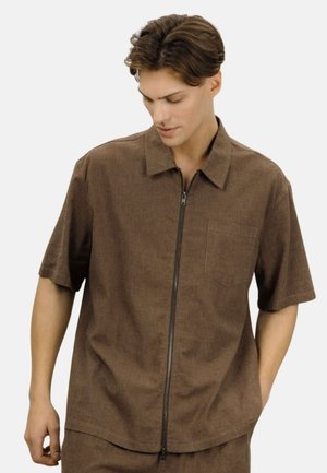 Ung mann iført en brun kortermet genser med glidelås og brystlomme, som ser ned med hendene avslappet langs siden.