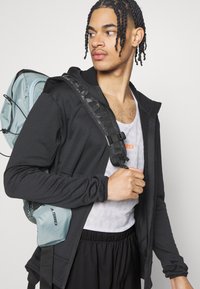 Sudadera negra de manga larga con cremallera en el frente, paneles de malla, llevada sobre una camiseta sin mangas blanca. Mochila azul claro con correas ajustables y logotipos.