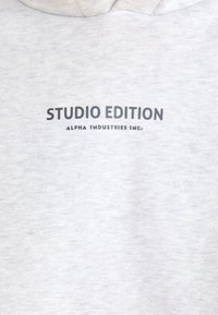 Haut de survêtement gris clair à texture douce, avec un texte imprimé noir : "ÉDITION STUDIO" et "ALPHA INDUSTRIES INC." à l'avant.