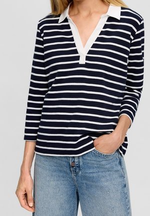 Woman wearing a navy and white striped long-sleeve V-neck top with white collar, paired with light blue denim jeans, one hand in pocket.