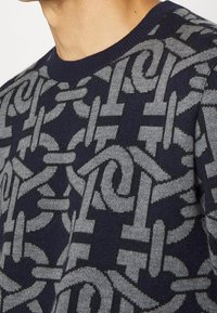 Maglione a maglia blu navy con un motivo a catena grigia ripetuto, collezione rotonda e tessuto testurizzato. La vista ravvicinata mostra i dettagli e il design.