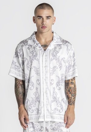 Młody mężczyzna z bardzo krótkimi włosami, ubrany w białą koszulę z krótkim rękawem, ozdobioną szarymi, ozdobnymi kwiatowymi wzorami, z widocznymi tatuażami na ramionach i klatce piersiowej.