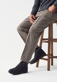 Cizme negre cu șireturi și finisaj texturat, pantaloni cargo bej cu ușoară strălucire, așezate pe un scaun de lemn pe un fundal alb simplu.