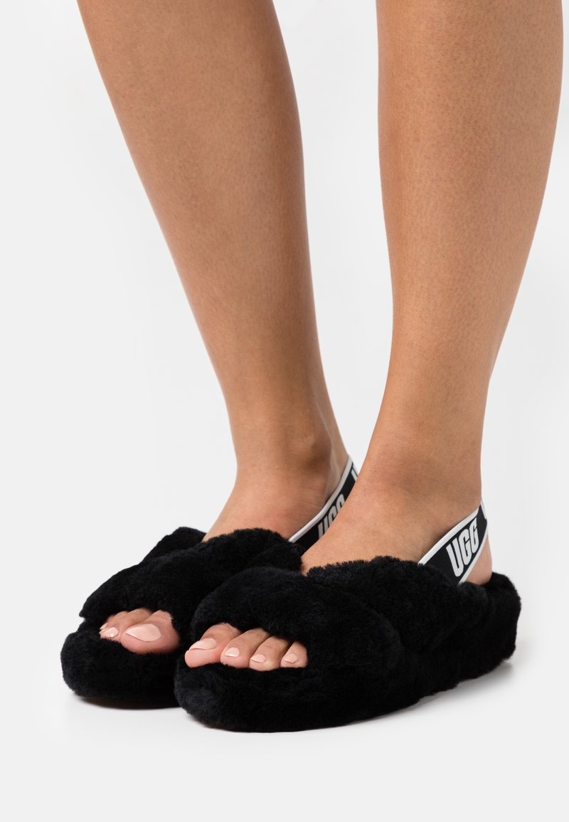 Pieds portant des chaussons noirs duveteux à bout ouvert avec des sangles élastiques marquées "UGG" au talon.