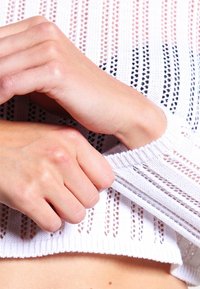 Pull en laine blanche avec un tricotage lâche et ouvert. Les poignets sont côtelés et des mains ajustent la manche. Diverses textures visibles.