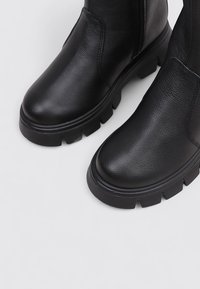 Botas de tobillo de cuero negro con suela gruesa, punta redondeada y superficie texturizada. Presenta un acabado suave y cremallera lateral para facilitar su uso.