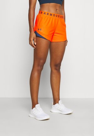 Žena má na sobě jasně oranžové sportovní šortky Under Armour, tmavě modrý sportovní top a bílé běžecké boty, stojí na šedé podlaze u bílé stěny.