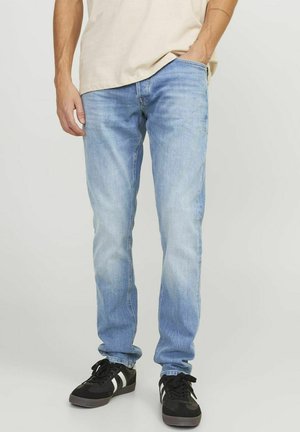 Mężczyzna w jasnoniebieskich obcisłych dżinsach i czarnych trampkach z białymi paskami, stojący z jedną ręką w kieszeni na jednolitym tle.