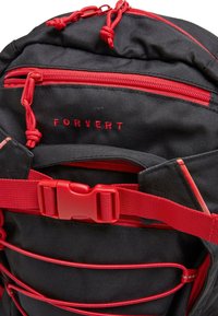 Juodas kuprinė su raudonais priedais, užtrauktukais ir reguliuojamomis petnešomis. Ypatybės: logotipas, virvės detalės ir tvirtas audinio tekstūra.