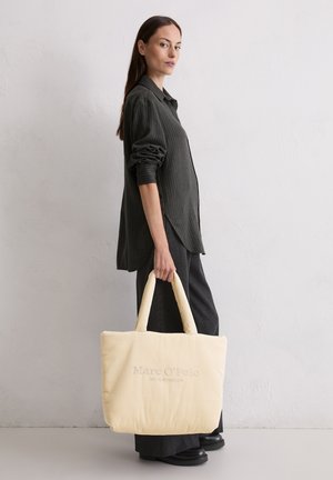 Beige Cordura Tasche mit zwei dicken Griffen, mit dem embossed Text "Marc O'Polo" auf der Vorderseite. Getragen mit einem dunklen Hemd und Hose.