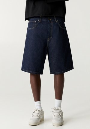 Shorts en denim bleu foncé portés avec des chaussettes blanches et des baskets de couleur claire, visibles de la taille aux pieds sur un fond uni.