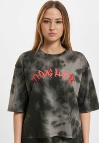 Šedé oversized tričko s motifem tie-dye vyrobené z bavlněné směsi. Má krátké rukávy a na přední straně červený grafický text ve stylizovaném písmu.