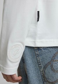 Weißes Langarmhemd mit strukturiertem Stoff, ausgestattet mit einem gestickten Logo am Ärmel und einem schwarzen Markenschild an der Seitennaht.