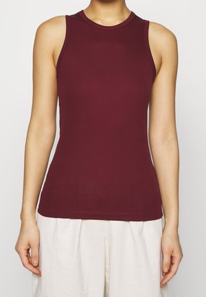 Vrouw draagt een mouwloze, geribbelde, bordeauxrode top gecombineerd met een wijde, witte broek met hoge taille, tegen een effen lichte achtergrond.