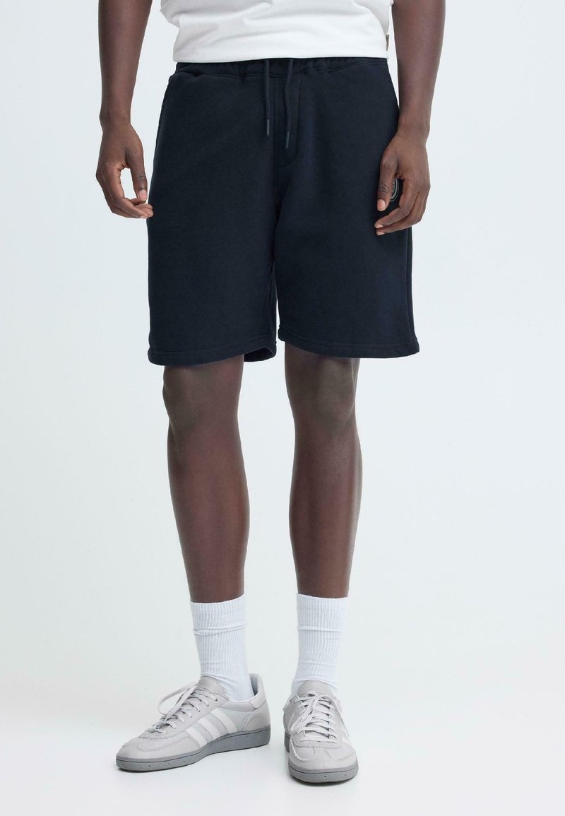 Shorts en coton noir avec une taille élastique et un cordon de serrage. Texture lisse, longueur au genou, assortis à des baskets grises et des chaussettes blanches.