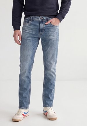 Mann trägt hellblaue ausgewaschene Jeans, dunkelblauen Pullover, weiße Sneakers mit roten und blauen Streifen, eine Hand in der Tasche.