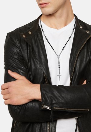 Chaqueta de cuero negra con cremalleras, camiseta blanca debajo y un collar largo de cuentas negras con un colgante de cruz de plata.