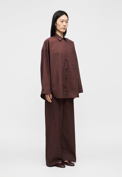 Femme debout portant une chemise ample brun foncé à boutons, un pantalon large assorti et des chaussures pointues sur un fond clair uni.