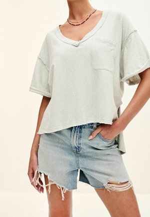 Kobieta ubrana w luźną, jasnoszarą koszulkę z dekoltem w serek i kieszenią z przodu oraz zniszczone jasnoniebieskie szorty dżinsowe z postrzępionymi krawędziami.