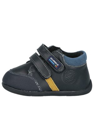 Zapato negro para niños con dos cintas de Velcro, acentos de ante azul marino, raya amarilla, puntera texturizada y suela de goma. Diseño redondeado.