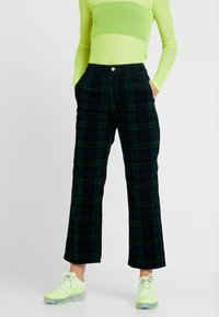 Personne portant un haut à manches longues translucide vert fluo, un pantalon à carreaux vert foncé et bleu marine, et des baskets vert fluo avec des semelles transparentes.