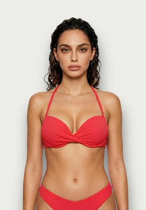 Kobieta o falowanych ciemnych włosach, ubrana w czerwony bikini z fakturą i wiązaniem na szyi, stojąca na jednolitym jasnym tle, patrząca prosto przed siebie.
