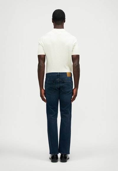 Homem em pé de costas, vestindo uma polo branca de mangas curtas, jeans azul escuro e sapatos sociais pretos, contra um fundo simples.